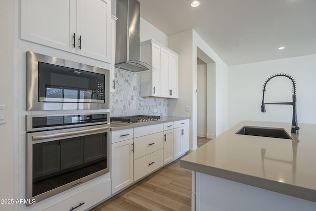 kitchen with sink, white cabinets, stainless steel appliances, wall chimney range hood, and light wood-type flooring