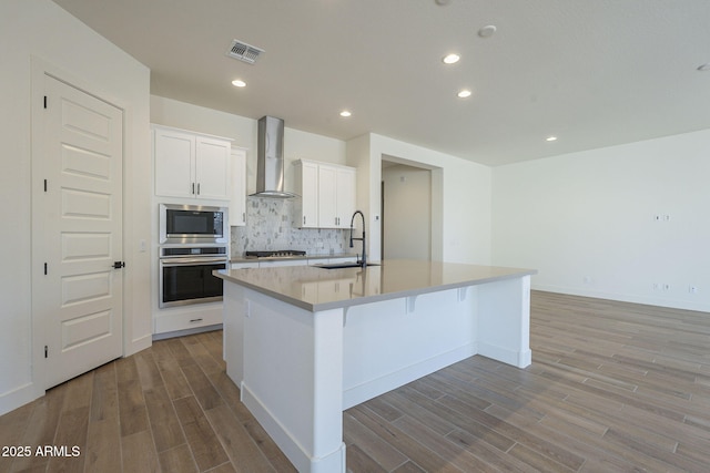 kitchen featuring sink, appliances with stainless steel finishes, wall chimney range hood, a kitchen island with sink, and white cabinets