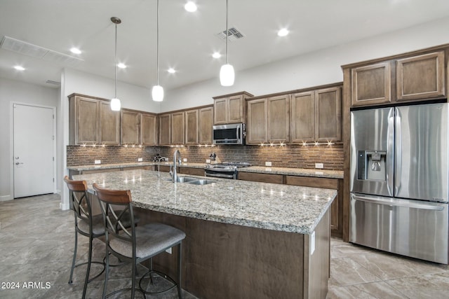 kitchen with a kitchen island with sink, sink, hanging light fixtures, appliances with stainless steel finishes, and light stone counters