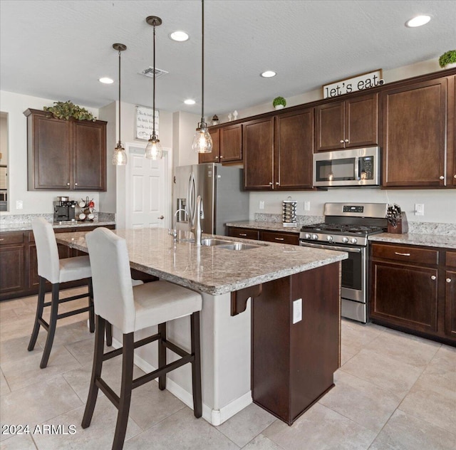 kitchen featuring an island with sink, light stone countertops, dark brown cabinetry, pendant lighting, and appliances with stainless steel finishes