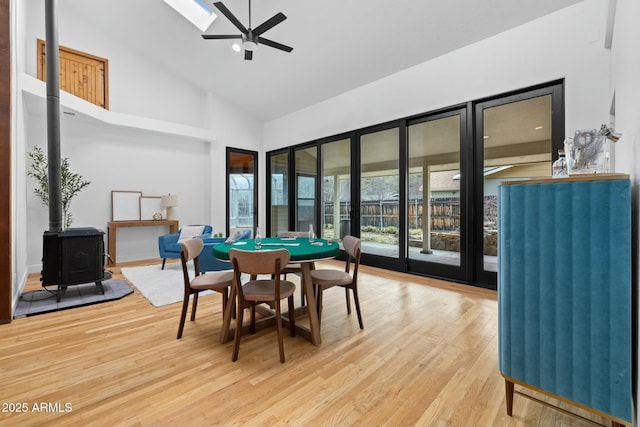 dining space with a skylight, light wood finished floors, a wood stove, ceiling fan, and high vaulted ceiling