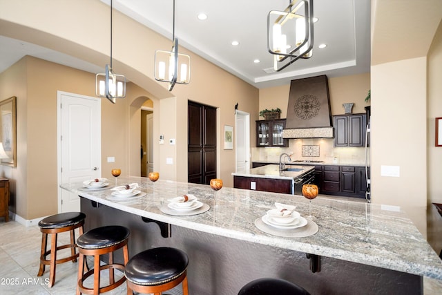 12904 W Dale Ln, Peoria, AZ 85383 - kitchen featuring premium range hood, arched walkways, an island with sink, dark brown cabinets, and a breakfast bar