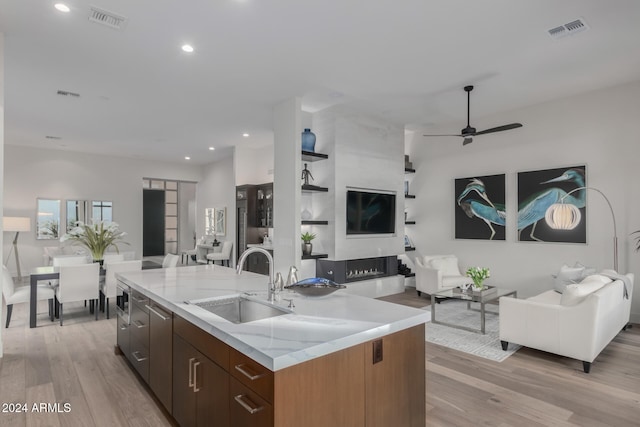 kitchen with a kitchen island with sink, sink, light wood-type flooring, light stone counters, and ceiling fan