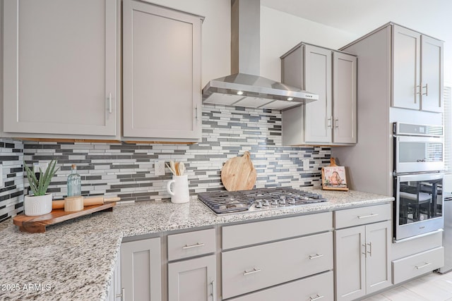 kitchen with gray cabinets, appliances with stainless steel finishes, decorative backsplash, light stone counters, and wall chimney exhaust hood