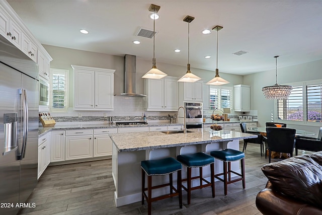 kitchen featuring white cabinets, wall chimney range hood, sink, built in appliances, and an island with sink