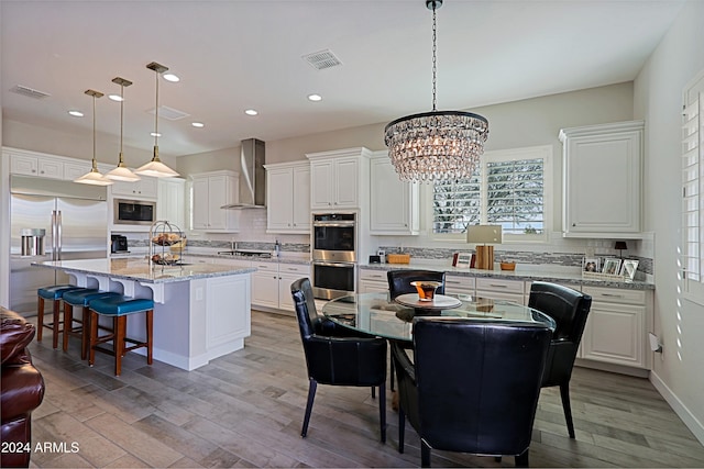 kitchen with a center island with sink, wall chimney exhaust hood, built in appliances, and hanging light fixtures