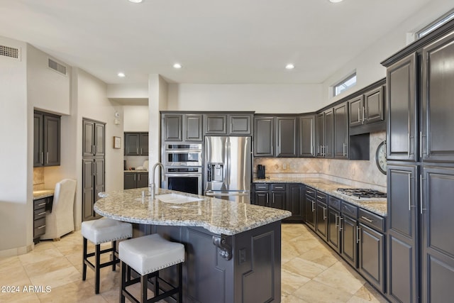 kitchen featuring sink, stainless steel appliances, light stone counters, decorative backsplash, and a center island with sink