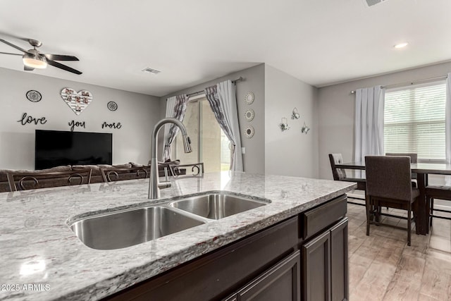 kitchen with a sink, dark brown cabinets, light stone counters, open floor plan, and a ceiling fan