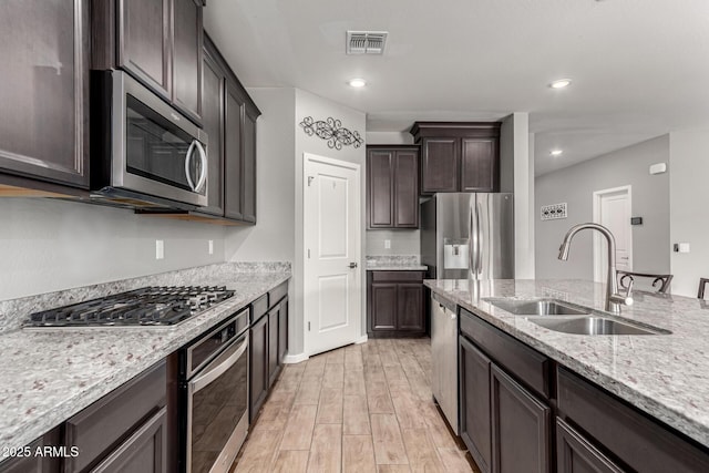 kitchen featuring appliances with stainless steel finishes, a sink, light wood finished floors, dark brown cabinetry, and light stone counters
