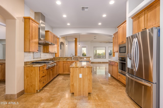 kitchen featuring light stone counters, kitchen peninsula, a kitchen island, wall chimney range hood, and appliances with stainless steel finishes