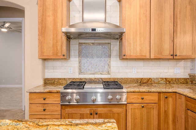 kitchen featuring ceiling fan, light stone counters, stainless steel gas cooktop, wall chimney exhaust hood, and backsplash