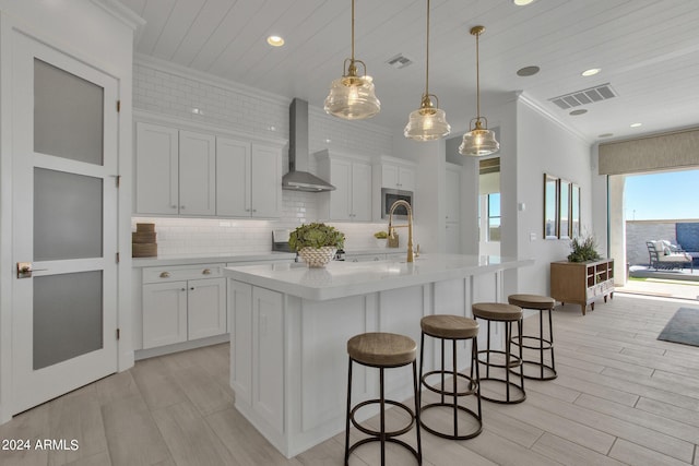 kitchen with white cabinets, decorative light fixtures, an island with sink, and wall chimney range hood