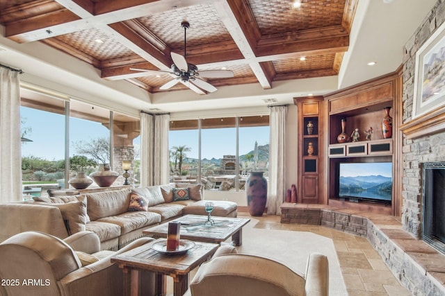 living room with stone tile flooring, ceiling fan, a stone fireplace, coffered ceiling, and beamed ceiling