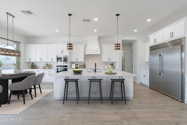 kitchen with sink, built in appliances, a kitchen island with sink, custom range hood, and white cabinets