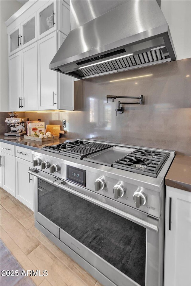 kitchen featuring stainless steel stove, white cabinets, glass insert cabinets, dark countertops, and wall chimney range hood