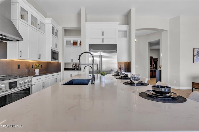 kitchen featuring backsplash, wall chimney range hood, built in appliances, light stone counters, and arched walkways