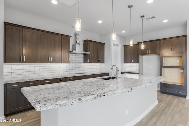 kitchen featuring an island with sink, decorative light fixtures, light stone countertops, light hardwood / wood-style flooring, and sink