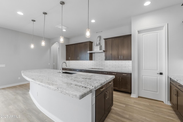 kitchen featuring backsplash, sink, light wood-type flooring, a kitchen island with sink, and dark brown cabinets