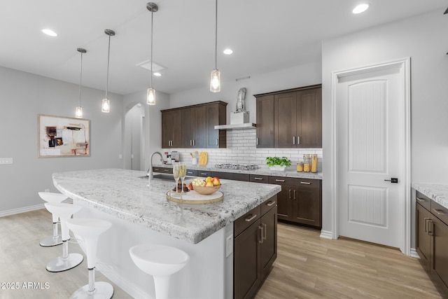 kitchen featuring light stone countertops, decorative light fixtures, tasteful backsplash, a kitchen island with sink, and dark brown cabinets