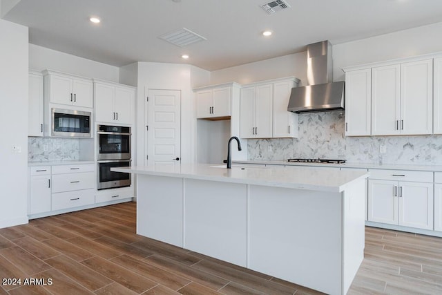 kitchen featuring appliances with stainless steel finishes, an island with sink, white cabinets, and wall chimney range hood