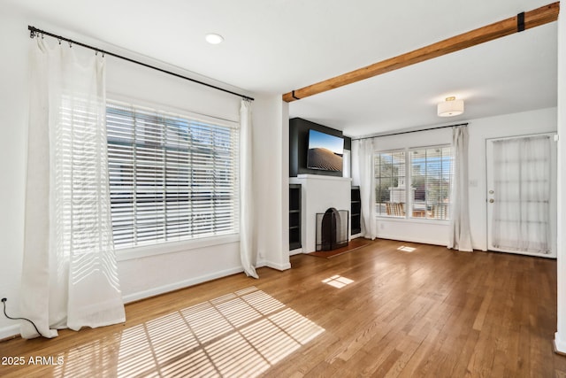 unfurnished living room featuring beam ceiling, a fireplace with raised hearth, baseboards, and wood finished floors