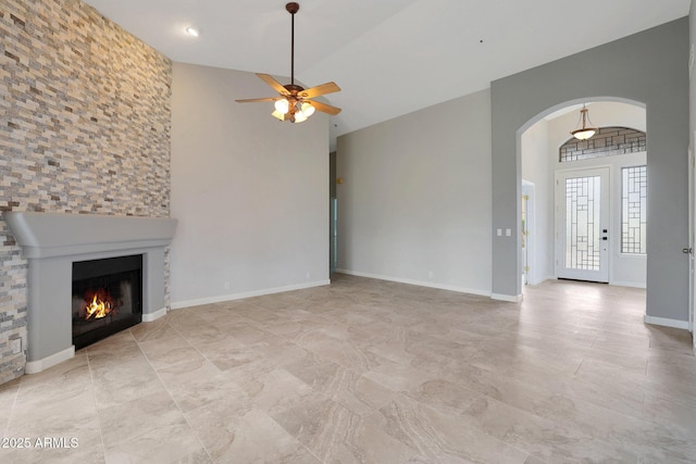 unfurnished living room with arched walkways, baseboards, a warm lit fireplace, ceiling fan, and high vaulted ceiling