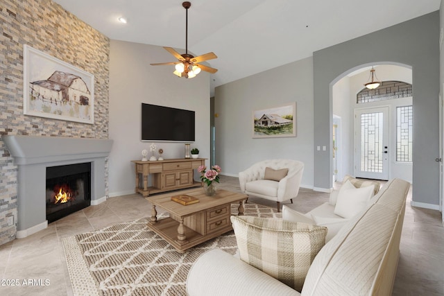 living area with high vaulted ceiling, a ceiling fan, arched walkways, a lit fireplace, and baseboards