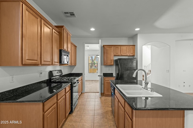 kitchen with black appliances, sink, light tile patterned flooring, an island with sink, and dark stone counters