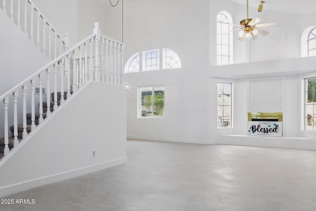 unfurnished living room featuring stairway, baseboards, a fireplace, a towering ceiling, and ceiling fan with notable chandelier