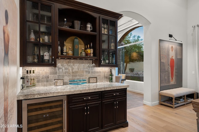 bar featuring wine cooler, tasteful backsplash, light stone countertops, light wood-type flooring, and dark brown cabinetry