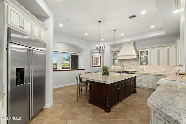 kitchen featuring tasteful backsplash, an island with sink, appliances with stainless steel finishes, dark brown cabinetry, and premium range hood