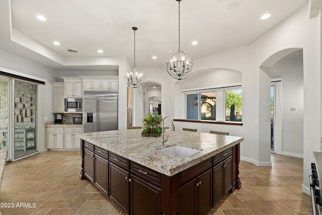 kitchen featuring a kitchen island with sink, built in appliances, a notable chandelier, sink, and backsplash