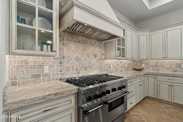 kitchen featuring backsplash, cream cabinetry, high end stainless steel range oven, custom range hood, and light stone countertops