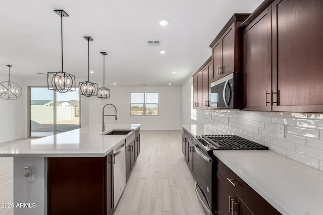 kitchen with sink, appliances with stainless steel finishes, a kitchen island with sink, backsplash, and decorative light fixtures