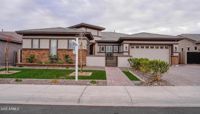 prairie-style house with brick siding, a fenced front yard, an attached garage, decorative driveway, and stucco siding