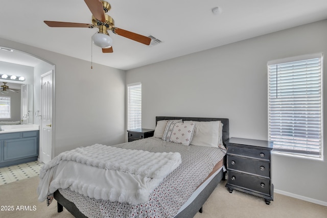 bedroom featuring baseboards, arched walkways, ceiling fan, ensuite bath, and light colored carpet