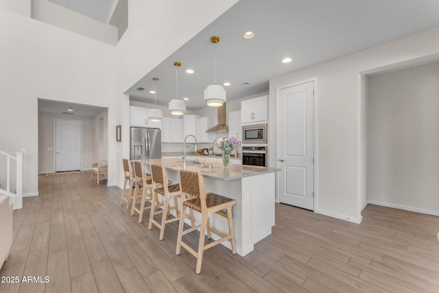 kitchen with wall chimney range hood, a kitchen island with sink, hanging light fixtures, stainless steel appliances, and white cabinets