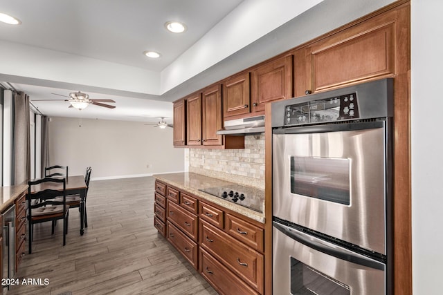 kitchen with black electric stovetop, backsplash, light stone counters, double oven, and hardwood / wood-style flooring