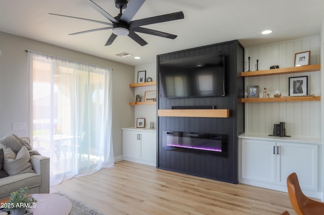 living area featuring light wood-type flooring, visible vents, a ceiling fan, and a glass covered fireplace