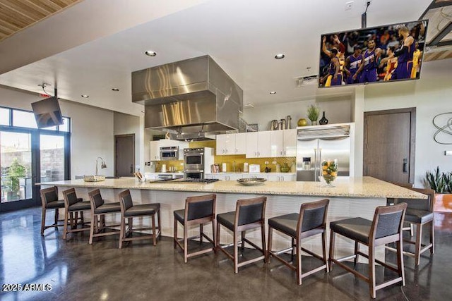 kitchen with a large island with sink, extractor fan, appliances with stainless steel finishes, white cabinetry, and a kitchen bar