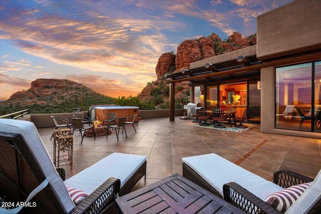 patio terrace at dusk with a mountain view, an outdoor living space with a fire pit, grilling area, and a hot tub