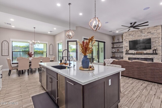 kitchen featuring ceiling fan with notable chandelier, sink, light hardwood / wood-style floors, a stone fireplace, and an island with sink