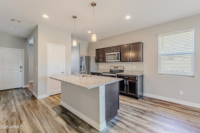 kitchen featuring an island with sink, appliances with stainless steel finishes, decorative light fixtures, light stone countertops, and dark brown cabinetry