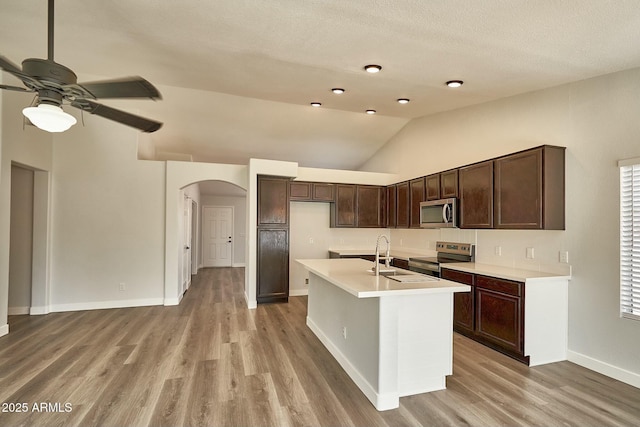 kitchen featuring arched walkways, an island with sink, appliances with stainless steel finishes, light countertops, and a sink