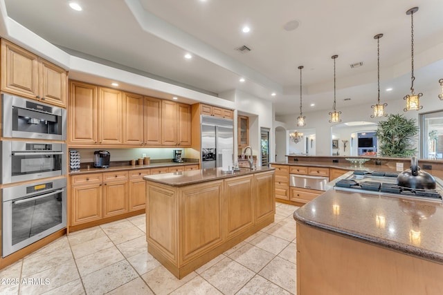 kitchen featuring appliances with stainless steel finishes, an island with sink, dark stone counters, hanging light fixtures, and a tray ceiling