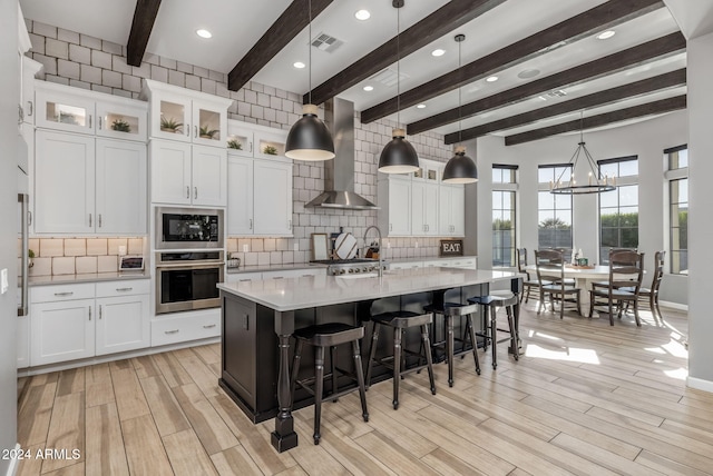 kitchen featuring wall chimney exhaust hood, white cabinetry, an island with sink, pendant lighting, and stainless steel appliances