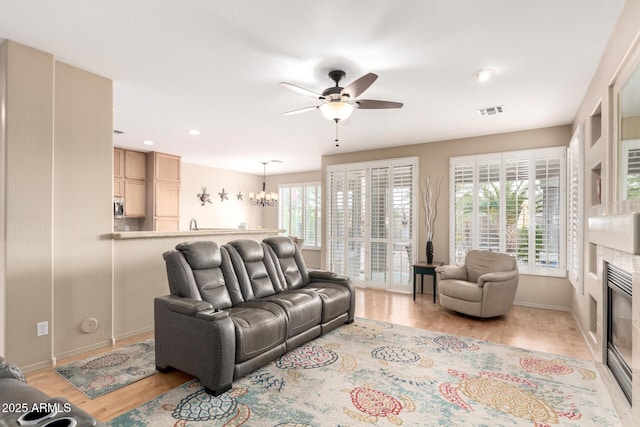 living room featuring ceiling fan with notable chandelier, visible vents, baseboards, light wood finished floors, and a glass covered fireplace