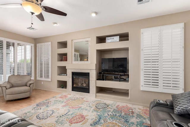 living area featuring built in shelves, wood finished floors, visible vents, a ceiling fan, and a tiled fireplace