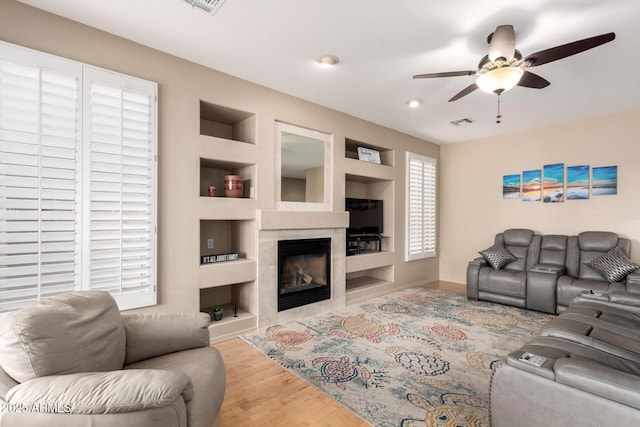 living room with built in shelves, a fireplace, visible vents, a ceiling fan, and wood finished floors