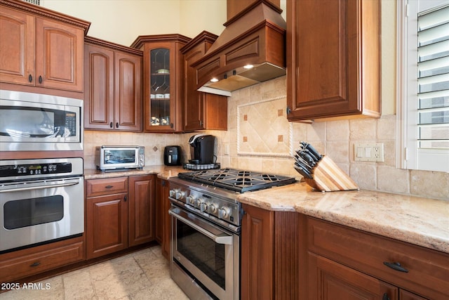 kitchen with light stone countertops, premium range hood, glass insert cabinets, appliances with stainless steel finishes, and backsplash